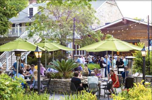 Outdoor dining scene with green umbrellas, people socializing, and lush greenery in a lively restaurant setting.