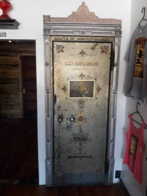An ornate, vintage bank vault door with intricate designs and a glass window, set against a wooden wall.