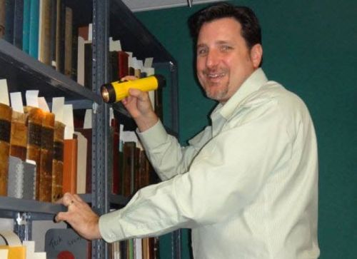 A man in a light-colored shirt smiles while holding a flashlight and examining books on a shelf.