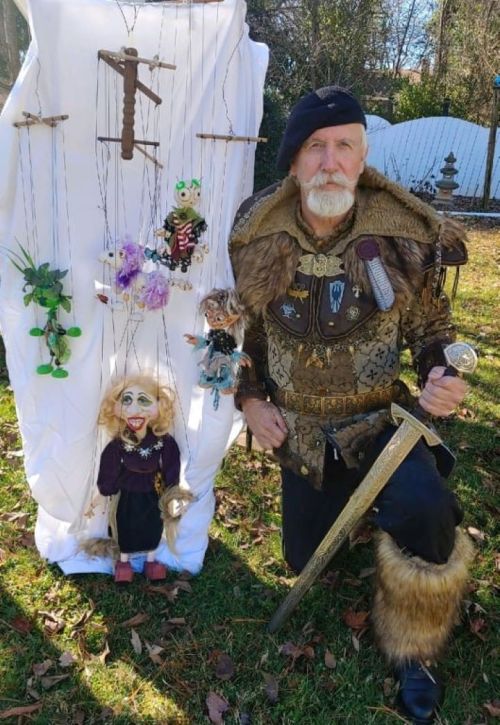 A man in a medieval costume poses with marionette puppets against a white backdrop in a garden setting.