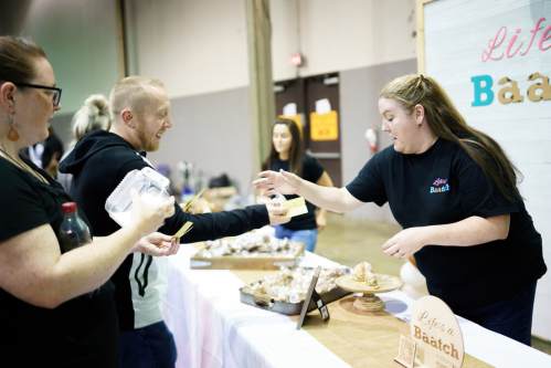 A vendor hands a sample to a customer at a market booth, with baked goods displayed on a table.