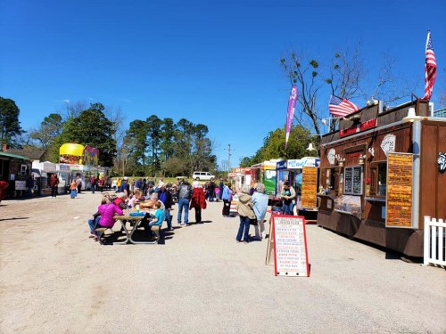 A bustling food truck area with people enjoying food, picnic tables, and colorful trucks under a clear blue sky.