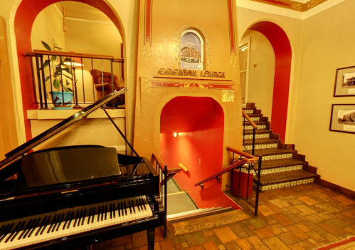 A grand entrance with a black piano, leading to a staircase and a sign for the Cocktail Lounge. Warm, inviting colors.