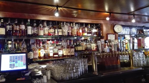 A well-stocked bar featuring various bottles of liquor, a clock, and a row of beer taps, with glasses in the foreground.