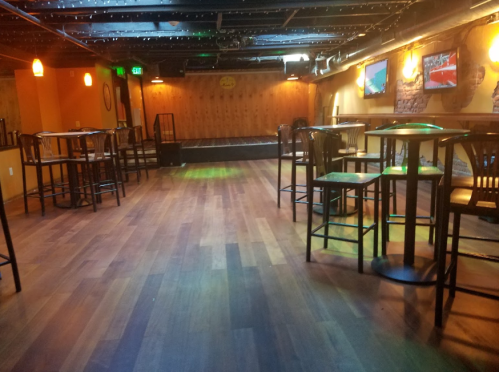 A spacious, empty bar with wooden floors, tall tables, and dim lighting, featuring a small stage in the background.
