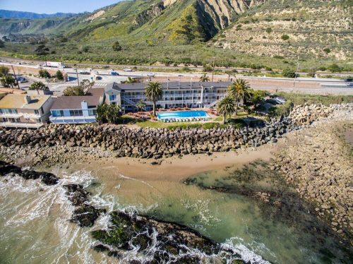 Aerial view of a coastal hotel with a pool, palm trees, and rocky shoreline, surrounded by hills and a highway.