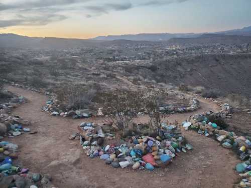 A winding dirt path lined with colorful stones, overlooking a valley at sunset with distant mountains.