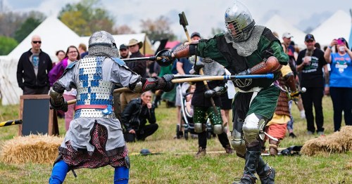 Two armored fighters engage in a mock battle with wooden weapons, surrounded by spectators and tents in the background.