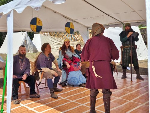 A group of people in medieval costumes seated under a tent, with one person standing and speaking to them.