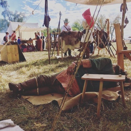 A reenactment scene with people in historical attire, one person resting on the ground, surrounded by tents and tools.
