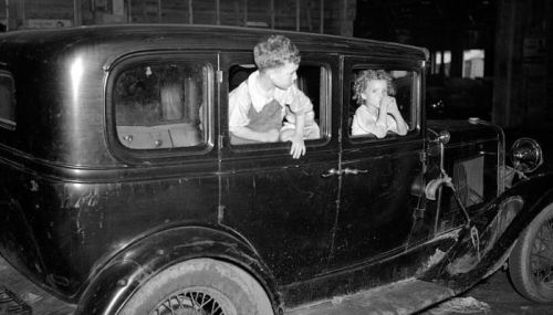 Two children peek out of the windows of an old black car in a dimly lit garage.