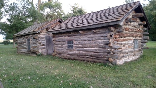 A rustic log cabin with a sloped roof, two windows, and a door, surrounded by green grass and trees.