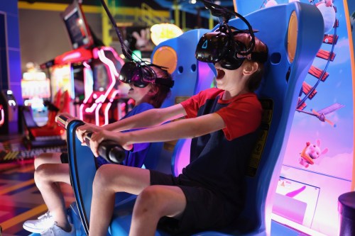 Two children wearing VR headsets sit in gaming chairs, excitedly experiencing a virtual reality game in an arcade.