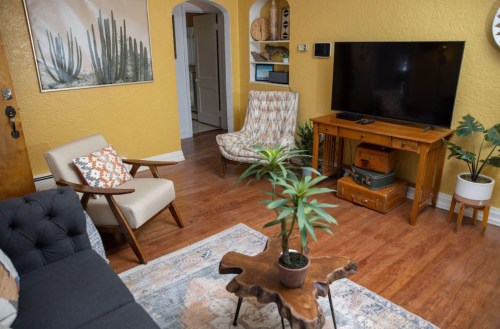 Cozy living room with yellow walls, a TV, plants, and stylish furniture, featuring a wooden coffee table and decorative art.