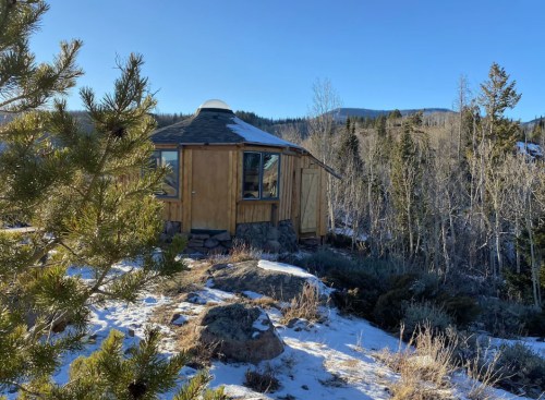 A wooden yurt surrounded by snow-covered ground and trees, with a clear blue sky in the background.