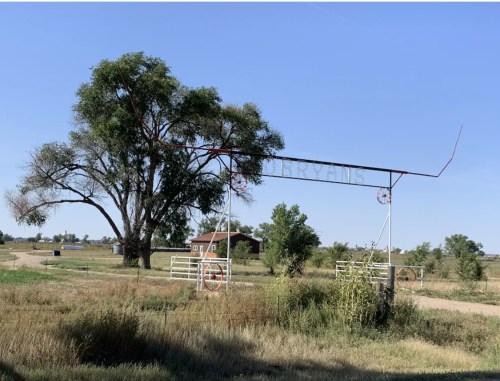 A rustic entrance gate with the word "Ranch" under a clear blue sky, surrounded by trees and open fields.