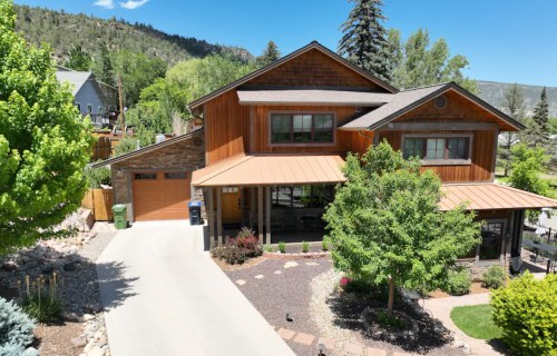 A modern two-story house with a wooden exterior, surrounded by trees and landscaped gardens on a sunny day.