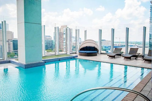 Rooftop pool with lounge chairs and a cozy cabana, surrounded by city skyline and blue sky.