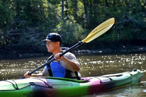 A person paddles a kayak on calm water, surrounded by trees, wearing a cap and a life jacket.