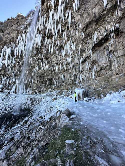 A person stands near a frozen waterfall, surrounded by icicles and icy terrain on a rocky cliffside.