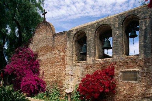 Historic brick wall with arched bell towers, adorned with vibrant pink and red flowers under a blue sky.