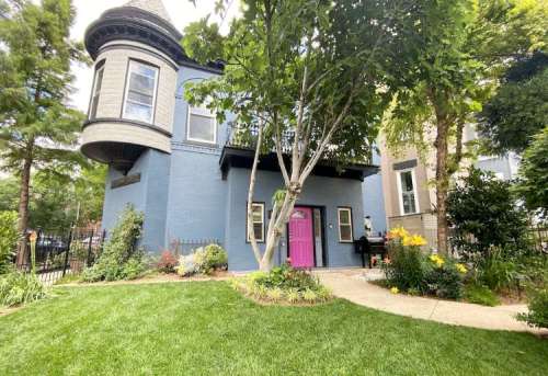 A charming blue house with a turret and a bright pink door, surrounded by greenery and flowers.