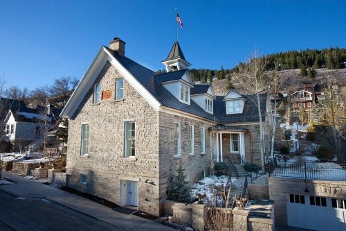A large stone house with a peaked roof and American flag, surrounded by snow and trees in a mountainous area.