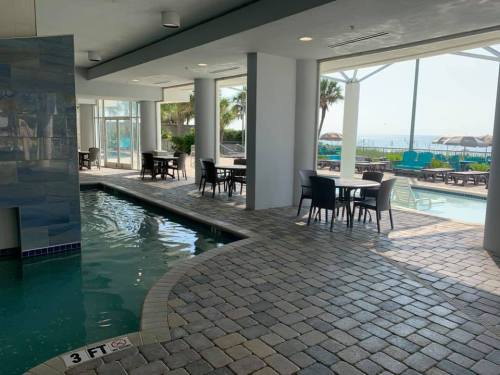 Indoor pool area with lounge chairs and tables, overlooking an outdoor pool and ocean view. Bright and inviting atmosphere.