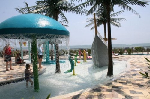 A water play area with palm trees, featuring a splash pad and children playing near the ocean.