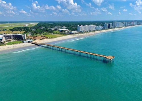 Aerial view of a long pier extending into turquoise waters, with a sandy beach and buildings in the background.