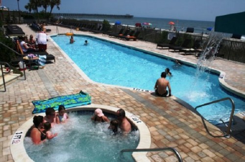 A sunny pool area with families enjoying the water, a hot tub, and a view of the ocean in the background.