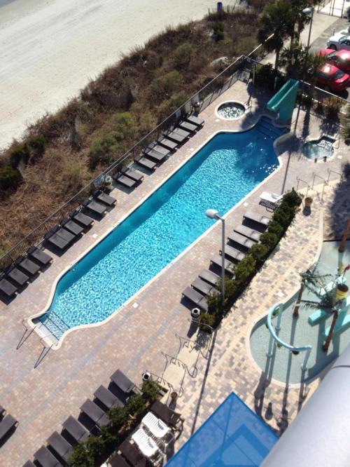 Aerial view of a blue swimming pool with lounge chairs, a slide, and a hot tub, surrounded by greenery and a beach.