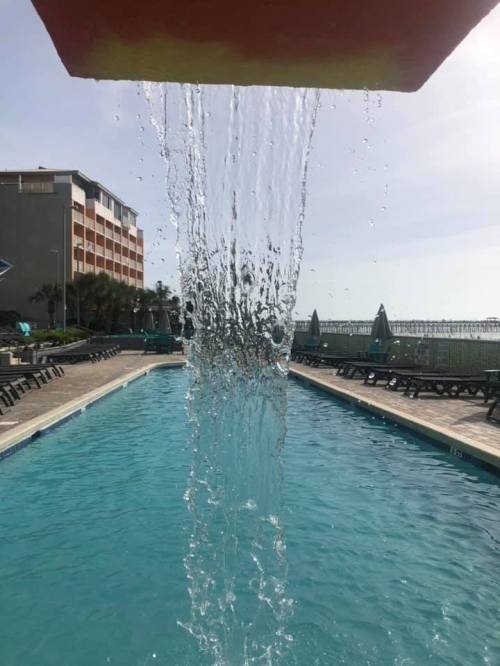 Water cascades from a colorful structure into a clear swimming pool, with lounge chairs and a pier in the background.