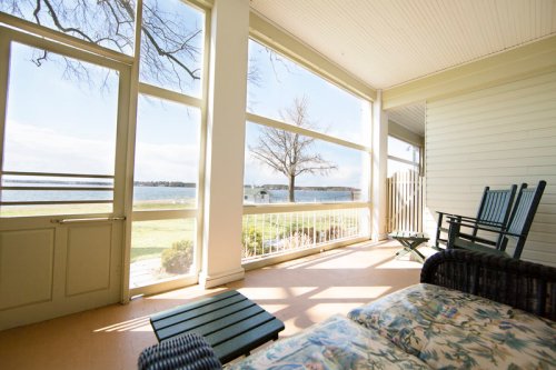 A serene porch with rocking chairs overlooking a lake, framed by large windows and a peaceful outdoor view.
