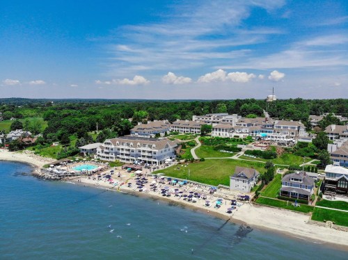 Aerial view of a beach resort with pools, green lawns, and guests enjoying the sandy shore under a blue sky.