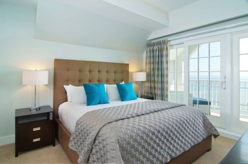 A cozy bedroom featuring a large bed with blue pillows, bedside tables, and a view of the ocean through glass doors.