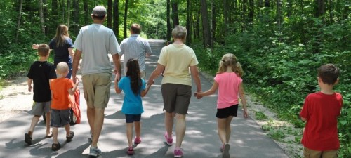A group of adults and children walking hand-in-hand on a forested path, surrounded by greenery.