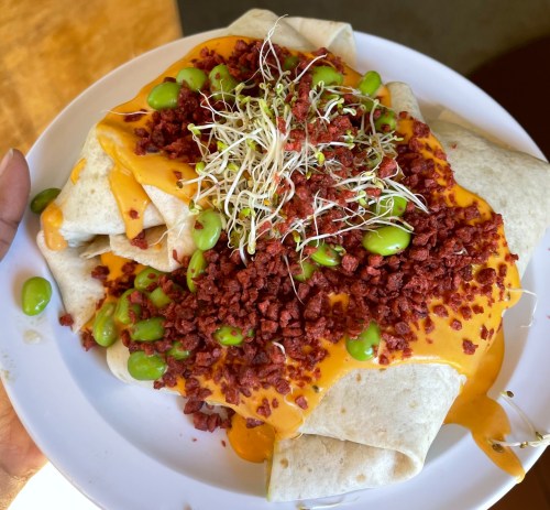 A plate of burritos topped with melted cheese, green edamame, and red chorizo, garnished with sprouts.