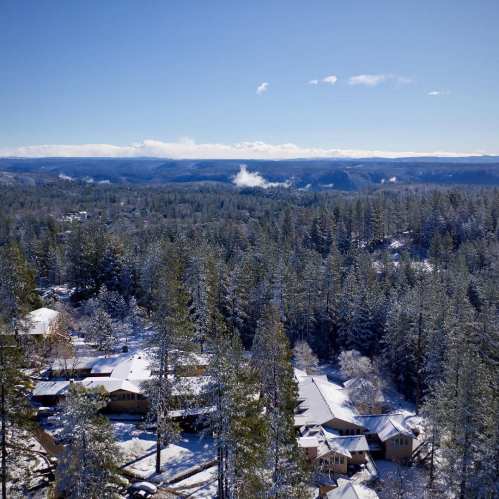 Aerial view of a snowy landscape with evergreen trees and houses nestled in a valley under a clear blue sky.