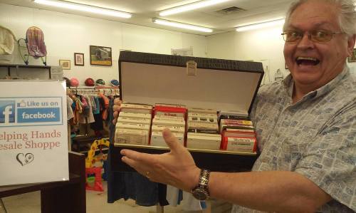 A smiling man holds a box of vintage cassette tapes in a thrift shop, with a sign promoting their Facebook page nearby.