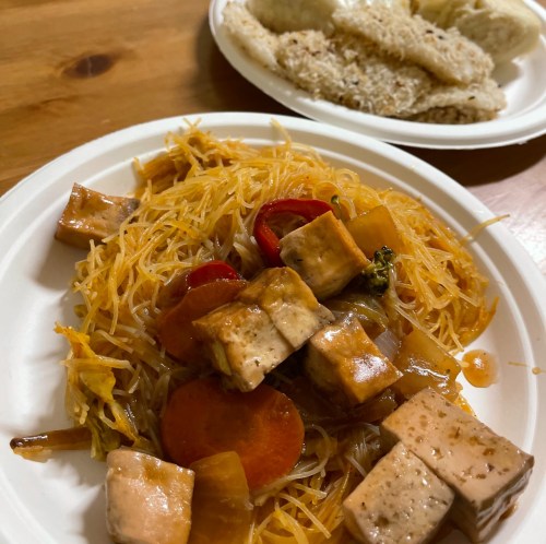 A plate of stir-fried noodles with vegetables and tofu, alongside a serving of rice cakes.