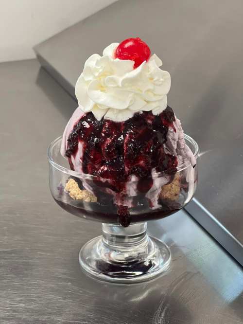 A glass dessert dish filled with purple ice cream, topped with whipped cream, a cherry, and berry sauce.