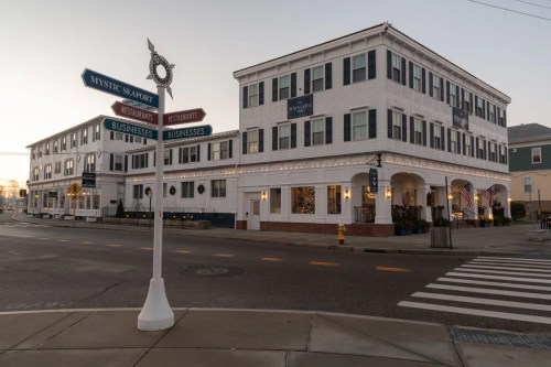 A street sign points to Mystic Seaport and nearby restaurants, with a charming white building in the background at dusk.