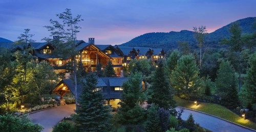 A scenic lodge surrounded by trees, illuminated at dusk with mountains in the background.