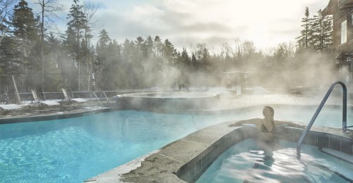 A woman relaxes in a steaming hot tub beside a tranquil pool surrounded by trees and misty air.