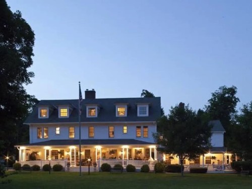 A large, elegant white house with multiple windows, illuminated at dusk, surrounded by trees and a lawn.