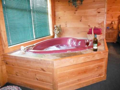 A cozy wooden bathroom featuring a pink jacuzzi tub, a glass of wine, and a bottle on the side, with green blinds.