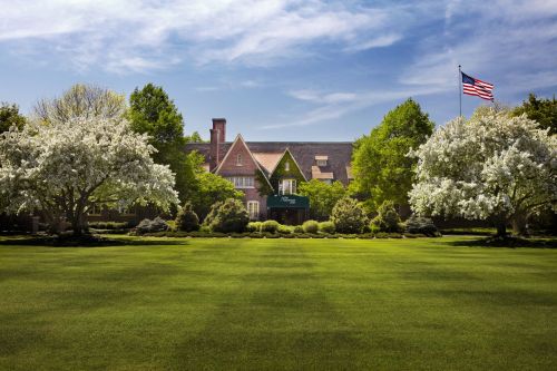A large, elegant building surrounded by lush green lawns and flowering trees, with an American flag in the background.