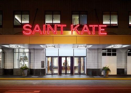 Neon sign reading "SAINT KATE" above the entrance of a modern building with large windows and potted plants.