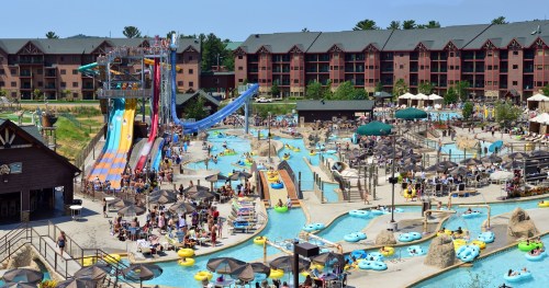A vibrant water park with slides, lazy rivers, and crowds enjoying the sun, surrounded by a hotel in the background.